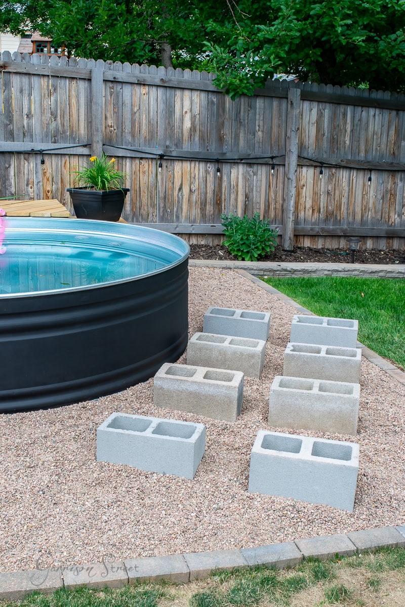 A round above-ground pool graces the outdoor space, framed by a wooden fence and trees. Set on gravel with cinder blocks around it, this area features a cozy sunbed and a plant in a black pot on the left.
