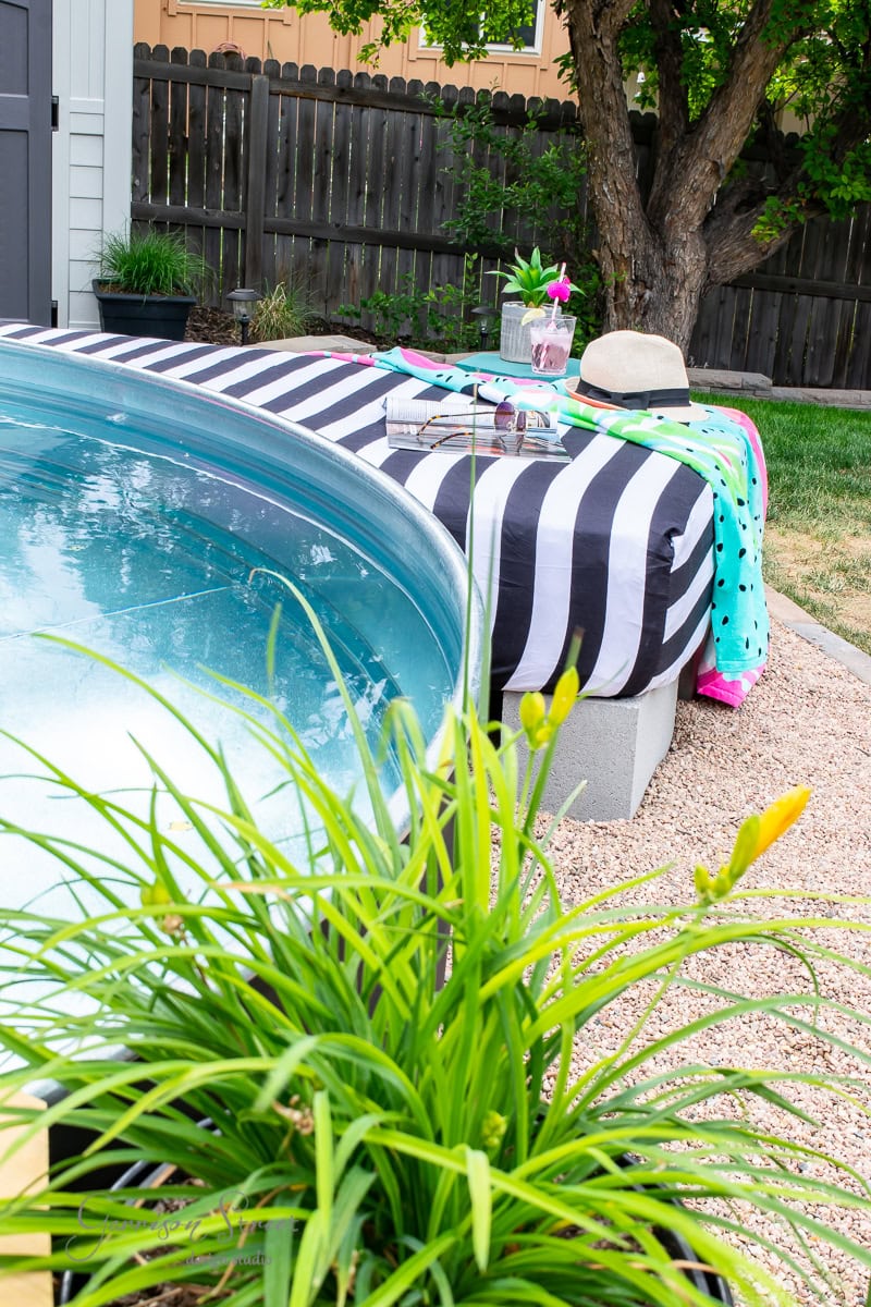 A small pool with a black and white striped cover sits beside a wooden fence. Nearby, a sunbed hosts a hat, a towel, and a drink on the side table. Green plants and pebbles frame the outdoor oasis, while a tree provides shade in the background.