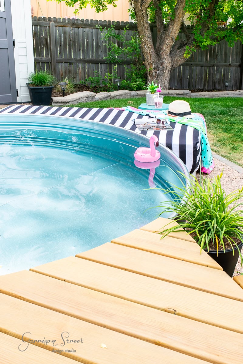 A small round pool with a wooden deck and an outdoor vibe is complemented by a black and white striped towel, sunglasses, and a drink nearby. A flamingo-shaped float bobs in the water. A sunbed rests beside potted plants, while a tree and fence provide a serene backdrop.