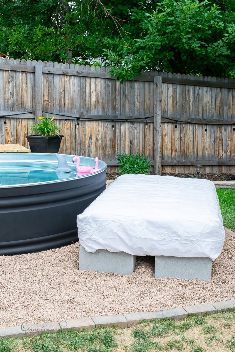 A round black stock tank pool with a pink inflatable flamingo sits beside a low rectangular bench draped with a white sheet. The area, surrounded by a wooden fence and lush greenery, offers an ideal sunbed retreat.