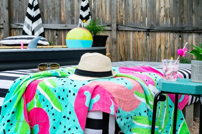 A poolside scene with a colorful towel featuring pink flamingos and green leaves. A straw hat and sunglasses rest on the chair. A striped inflatable tube and a beach ball add a summery vibe. A drink with ice and pink flamingo stirrer sits on a green table.