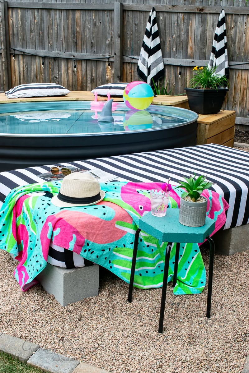 A backyard setup with a round above-ground pool, black and white striped cushions, and a table. A colorful towel, sunhat, and sunglasses rest on the table. Nearby, a small green side table holds a drink and a potted plant. Two plastic flamingos float in the pool beside the DIY sunbed arrangement.