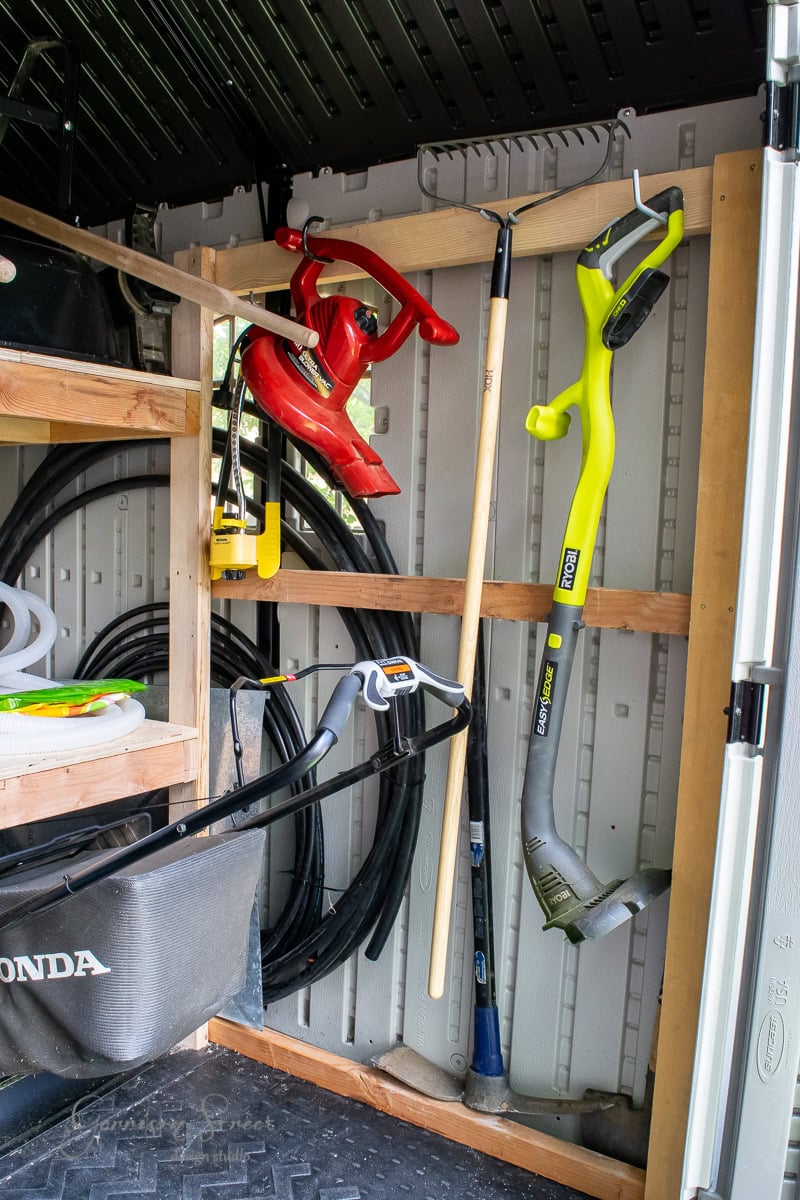 Inside a neatly organized shed, various garden tools hang on a wooden rack. A yellow string trimmer and a red leaf blower are visible. Coiled hoses and a lawnmower bag are neatly stored. DIY enthusiasts will appreciate how shelving units hold additional garden supplies efficiently.