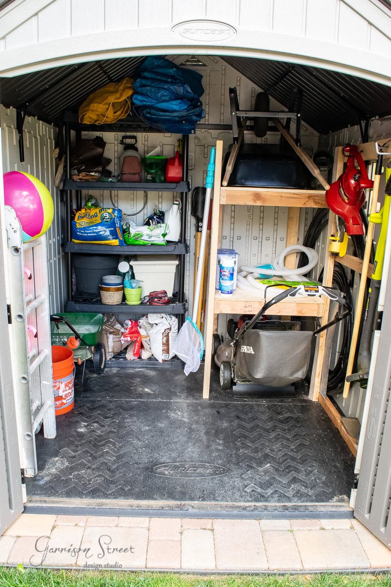 A neatly organized shed showcases DIY brilliance with shelves holding gardening tools, a lawnmower, a vibrant beach ball, bags of soil, a folded chair, and various supplies. The open doors reveal the tidy storage space accented by a practical rubber mat on the floor.
