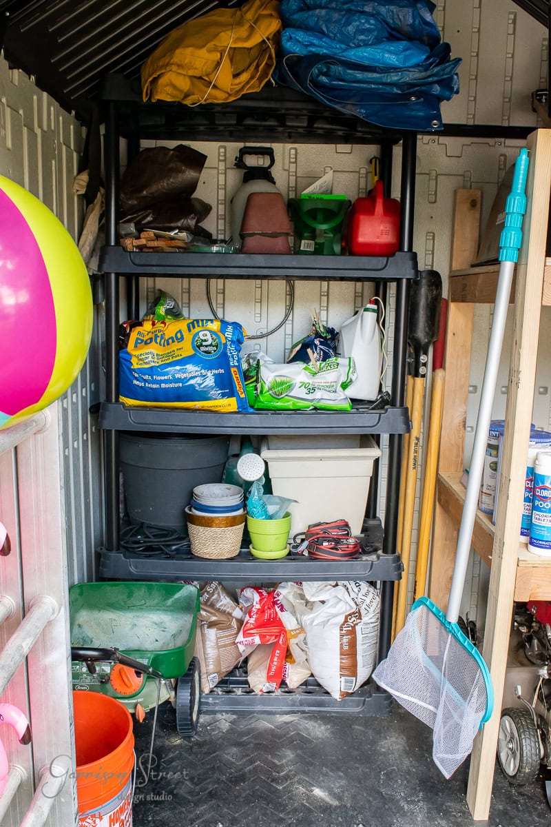 In a well-organized shed, shelves brim with gardening supplies like bags of potting soil, a watering can, and various tools. Nearby, a beach ball and lawn equipment await, while a fishing net leans against the wall, embodying DIY spirit with its neatly arranged chaos.