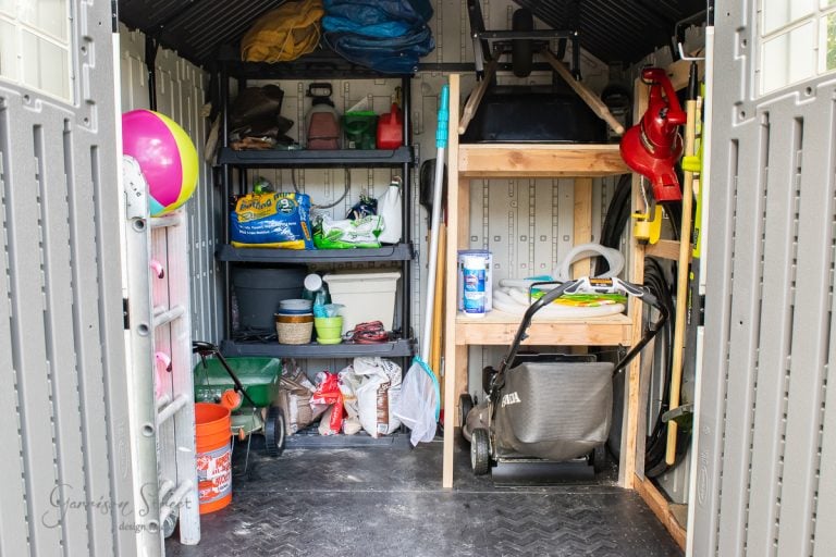 Interior of a cluttered storage shed with shelves holding various gardening tools, bags of soil, a lawn mower, a hose, and colorful balls. A ladder leans against the left wall, and a wheelbarrow is on the right.