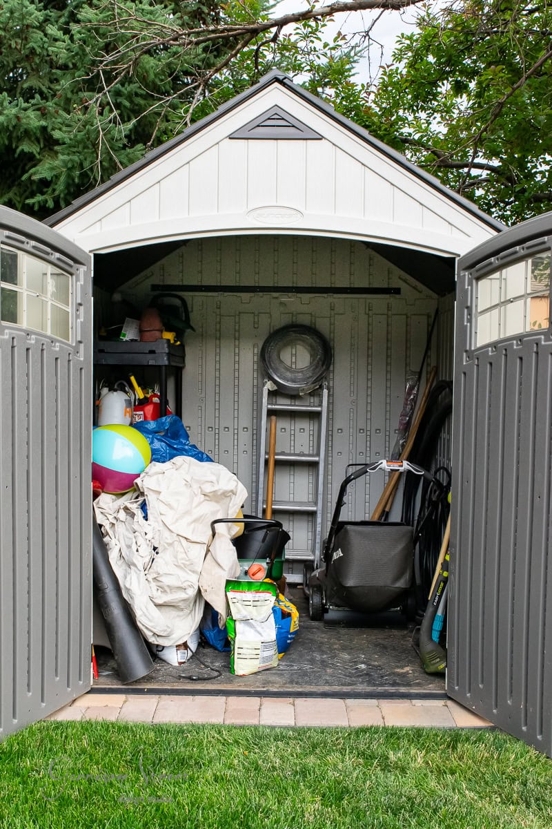 An open garden shed, carefully organized with DIY flair, is filled with items like a colorful beach ball, bags, a wheelbarrow, and garden tools. The shed harmonizes beautifully with the surrounding grass and trees.