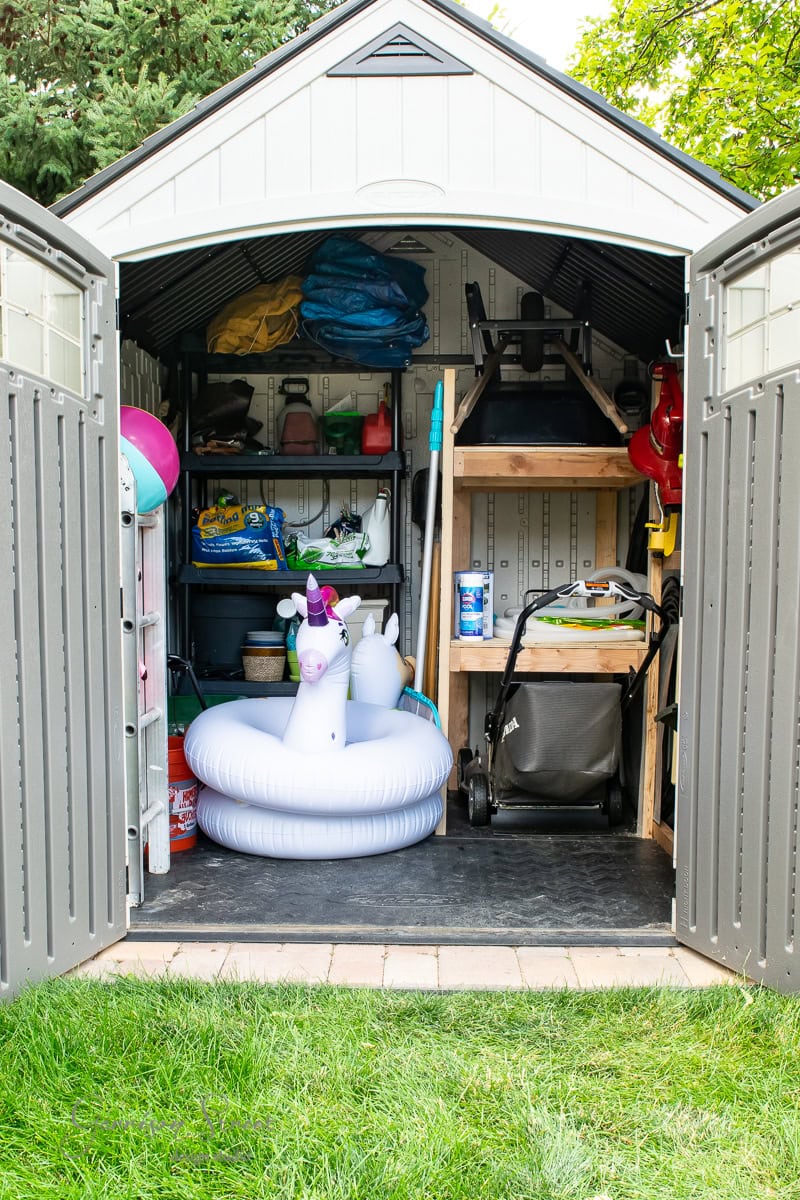 The open shed reveals a medley of items, from inflatable pool toys and a stroller to lawn equipment and tools, all neatly organized alongside storage bins. With grass and trees visible outside, this DIY haven blends practicality with the serenity of nature.