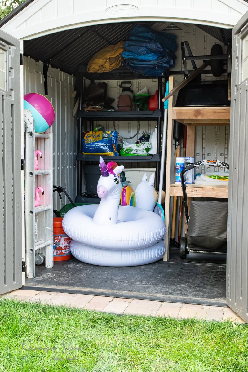 The open doors of the cluttered shed reveal a DIY project in need of organization, where garden tools, inflatable unicorn pool floats, and a beach ball lie amid various supplies on shelves and a wooden stand. The scene unfolds against the backdrop of green grass.