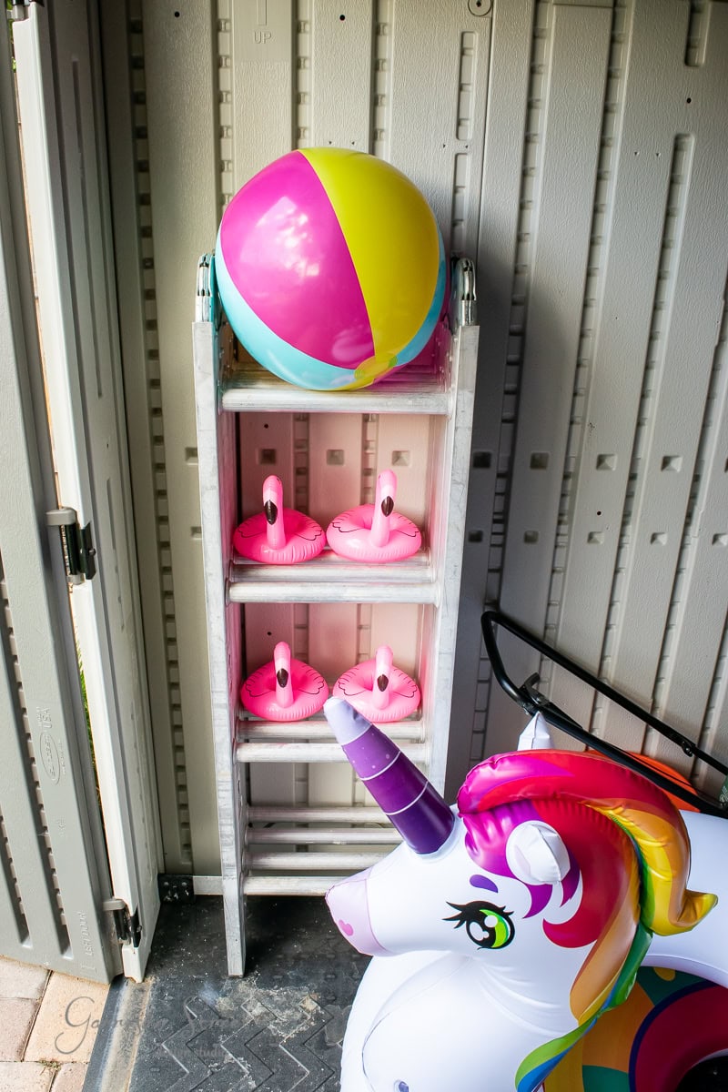 Bright and summery, the shed is a treasure trove of colorful pool toys, with a beach ball and unicorn float ready for action. Pink flamingo drink holders are meticulously placed on each step of a small ladder, showcasing impeccable DIY organization skills.