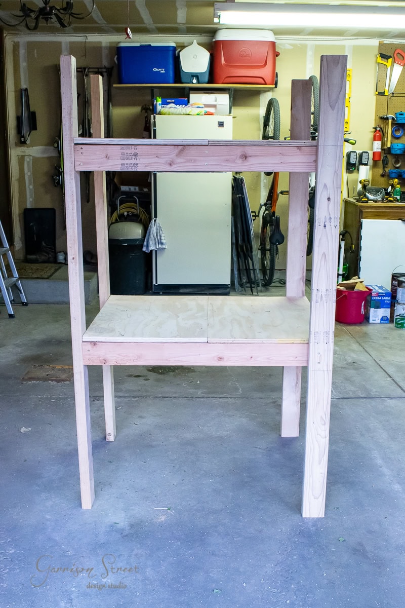 A wooden storage shelf is situated in a garage, perfect for DIY enthusiasts. It has two shelves made of light wood, ideal for organization. Various tools and items are visible in the background, including a ladder, refrigerator, and bicycles.