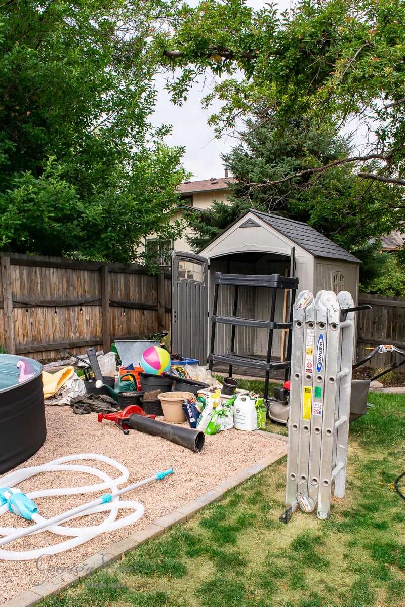 A cluttered backyard features various garden tools, hoses, a ladder, and a beach ball scattered on gravel. The storage shed hints at the need for organization, nestled among trees and bordered by a wooden fence. A hint of the swimming pool edge peeks out on the left.