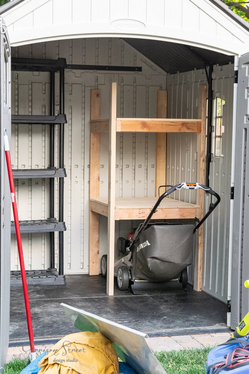 Inside the organized shed, a black plastic shelf on the left and a wooden shelf holding the lawn mower showcase DIY efficiency. The floor is covered in black material, while a broom rests against the wall amidst neatly arranged gardening tools.