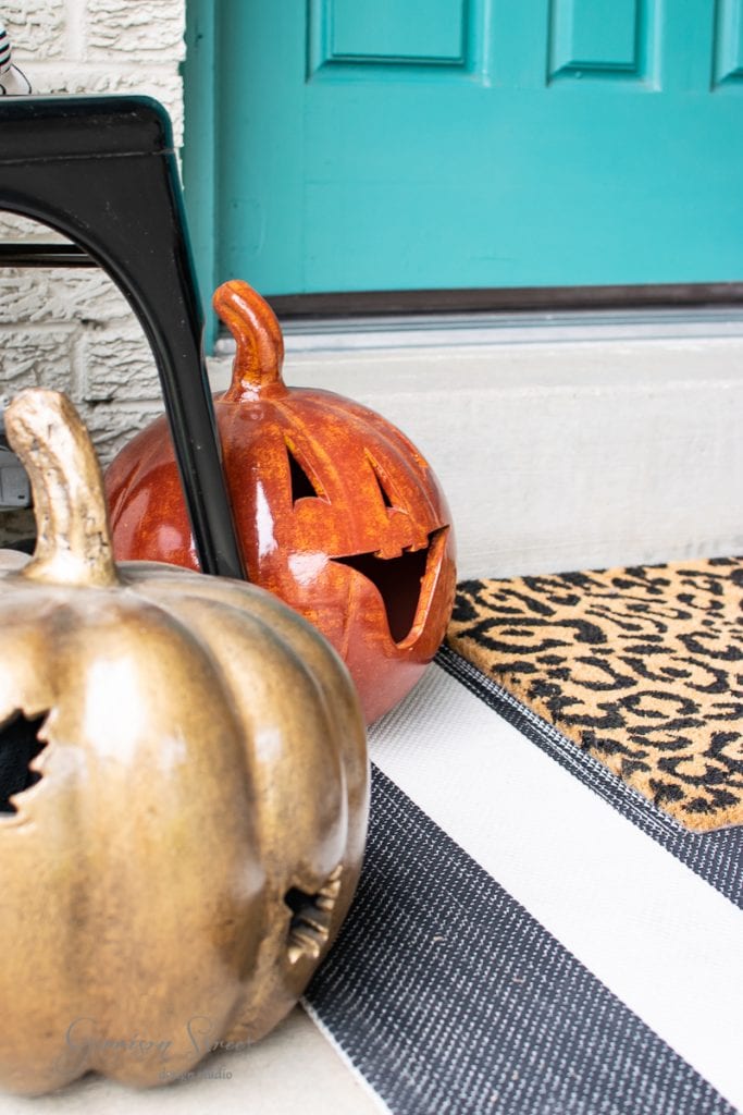 Home Two ceramic jack-o'-lantern pumpkins, one orange and one gold, sit on a striped mat by a teal porch door. This fall decor is completed with a doormat featuring a leopard print pattern.