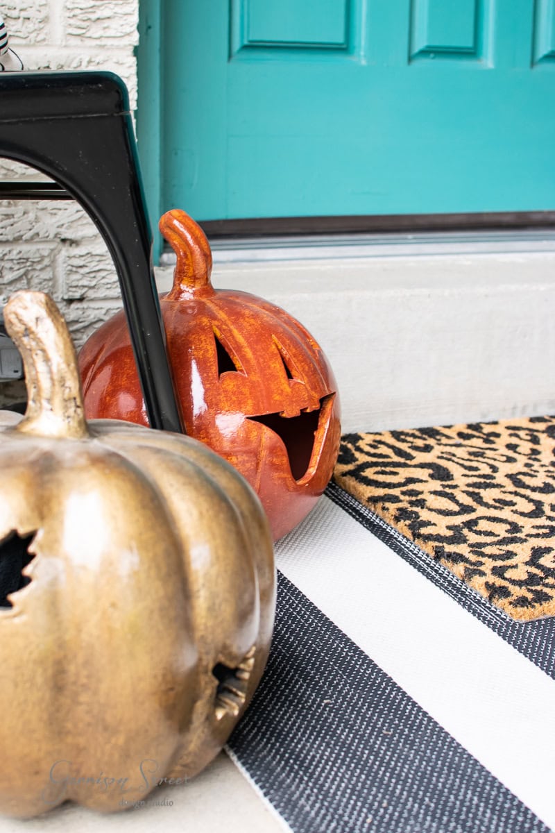 Two ceramic jack-o'-lantern pumpkins, one orange and one gold, sit on a striped mat by a teal porch door. This fall decor is completed with a doormat featuring a leopard print pattern.