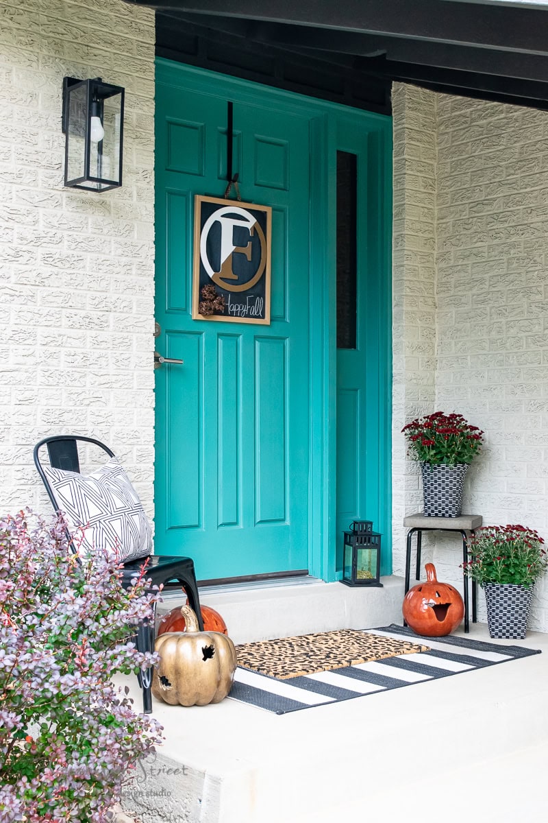 A turquoise front door with a monogram decor hangs under a black lantern. Pumpkins and chrysanthemums in pots embellish the fall porch, along with a striped doormat and a patterned chair, creating a cozy autumn scene.