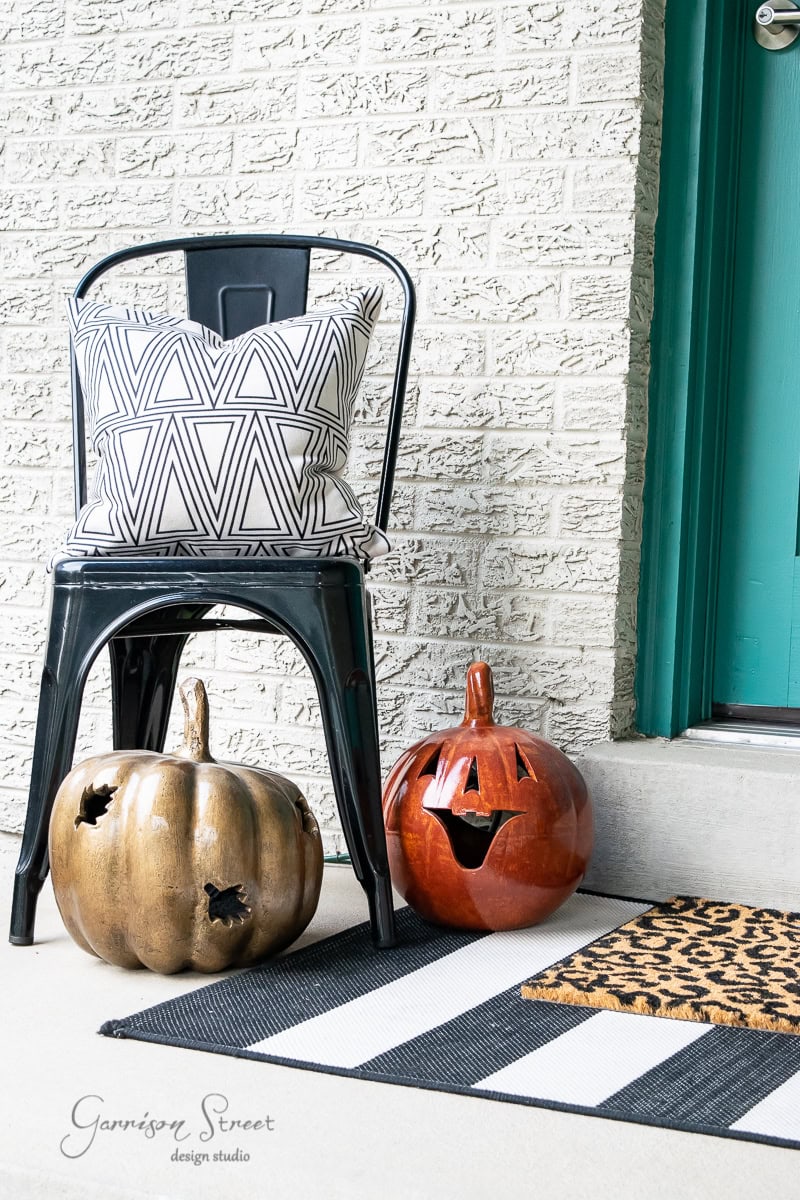 A black chair with a geometric patterned pillow graces a striped doormat on the porch, perfectly complementing the turquoise door. Beside the chair, embodying fall decor, are two decorative pumpkins—one bronze and one glossy orange with a carved face.
