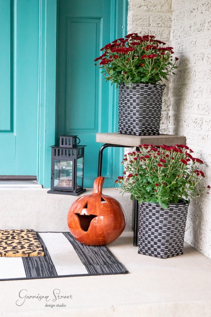 About A porch adorned with autumn decor: a carved pumpkin and two pots of red flowers set the scene, complemented by a black lantern and welcoming doormat. The teal door contrasts beautifully with the light-colored brick walls, creating a stunning fall ambiance.