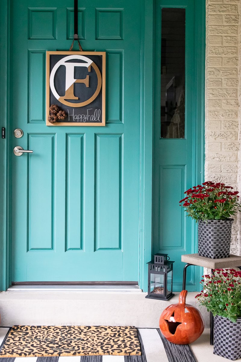 A teal front door showcases fall decor with a seasonal wreath. A doormat featuring animal print complements the porch, which is adorned with a carved pumpkin, a potted mum plant, and a black lantern. The wreath boasts an initial "F" alongside the cheerful greeting "Happy Fall.