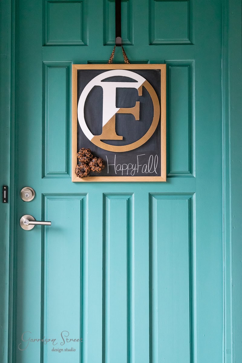 A teal door serves as the perfect backdrop for this fall decor, featuring a square frame with a decorative letter "F" in gold and white. The words "Happy Fall" are elegantly scripted underneath, while a small leopard print bow adds flair to this charming porch display.