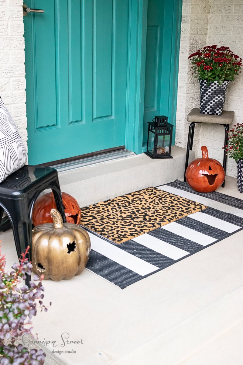 A festive fall porch with a teal door showcases a striped and leopard print doormat. Two carved pumpkin lanterns, one orange and one gold, grace either side. A black stool and potted plants complete the autumn decor, creating an inviting seasonal sanctuary.