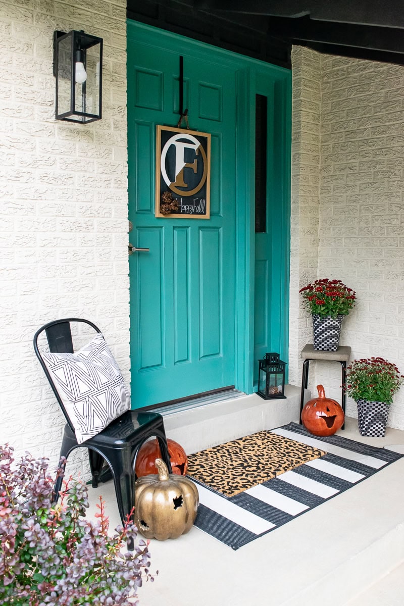 Front porch decor features a teal door adorned with pumpkins, potted plants, and a striped mat. A black chair with a geometric pillow sits near a lantern. A wooden sign with the letter "C" and "happy fall" warmly welcomes visitors.