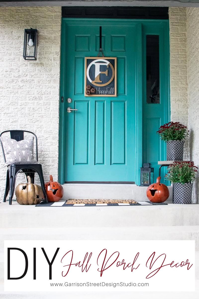 A teal front door flaunts a fall-themed wreath showcasing the letter "E." The porch, adorned with pumpkins—two artfully carved—and a black chair, captures the essence of autumn decor. Plants in baskets enhance this cozy fall setting, while a "DIY Fall Porch Decor" sign completes the charming scene.