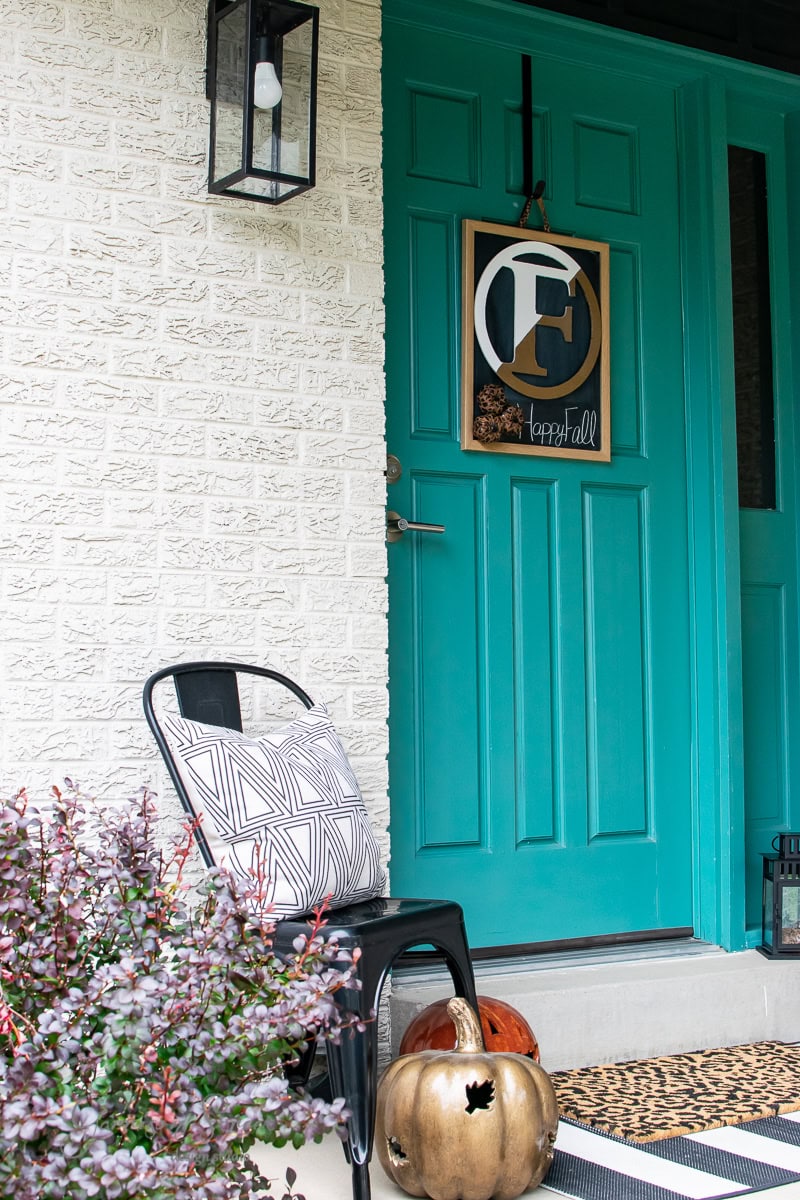 A teal front door welcomes autumn with a fall-themed wreath displaying the letter "E" and "happyfall." On the porch, a black metal chair with a geometric cushion sits next to a metallic pumpkin decoration. A small bush is in the foreground, completing the seasonal decor.