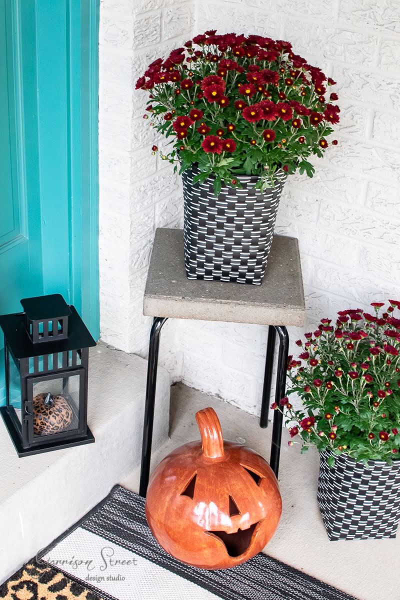 A welcoming porch displays fall decor with a jack-o'-lantern, a black lantern with a candle, and two pots of red chrysanthemums on a small table. The white brick wall and teal door add a vibrant backdrop to the cozy autumn scene.