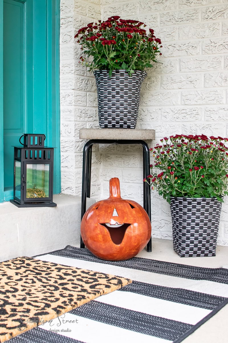 A fall porch decor includes a carved pumpkin, two pots of red chrysanthemums, a black lantern, and a striped doormat. The autumn setup is framed by a white brick wall and a teal door.