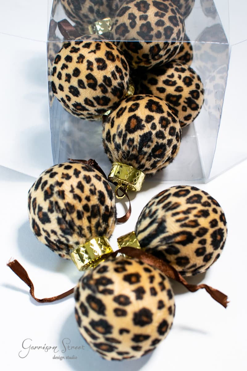 Christmas ornaments with a leopard print pattern are displayed, some inside a clear box and others outside. Perfect for a fall-inspired DIY project, the ornaments feature gold caps and brown hanging loops, adding a unique touch to your holiday decor or wreath arrangement.