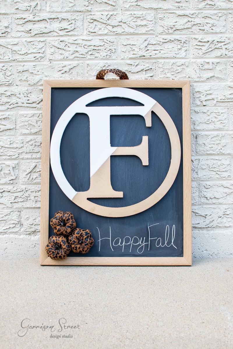 A wooden framed chalkboard featuring the letter "F" in white, encircled by a wreath-like design. Three decorative pinecones add a DIY touch. The words "Happy Fall" are elegantly scripted in white cursive at the bottom. It all rests against a charming white brick wall backdrop.