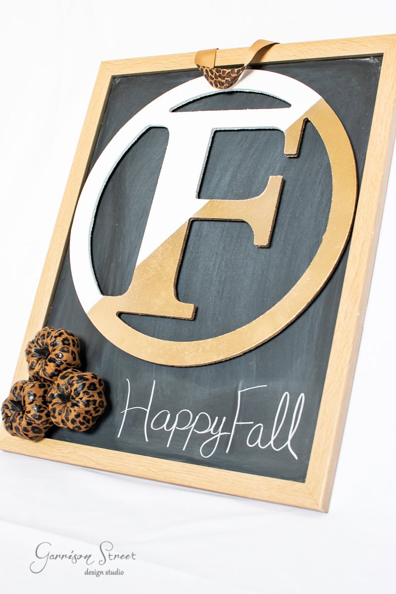 A wooden-framed chalkboard with the letter "F" in gold and white reads "Happy Fall" in elegant script. Decorative leopard print fabric pumpkins add a festive touch to this DIY creation, perfect as an autumn wreath alternative.