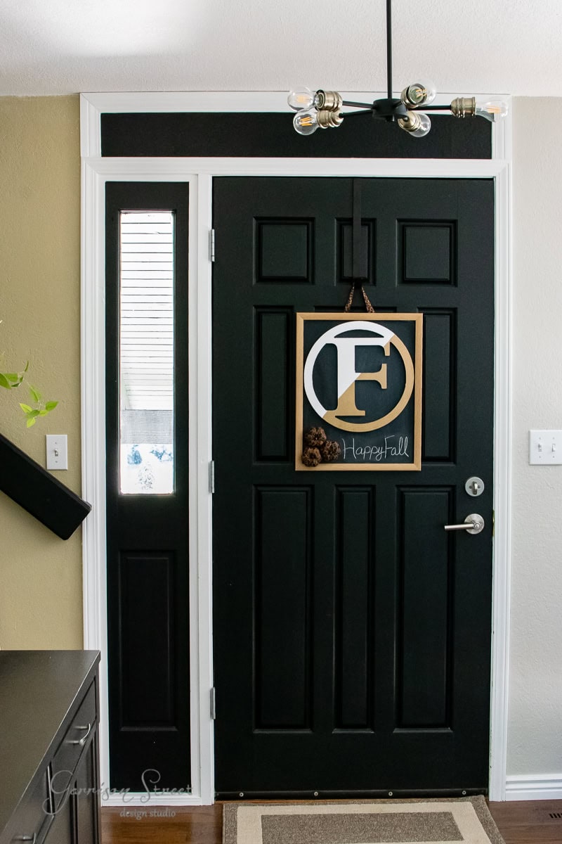 A black front door boasts a DIY touch with a vertical window on its left. A square wooden fall wreath, adorned with pinecones and a large letter "F," adds charm, while a small "Happy Fall" sign nests beneath it. Overhead, a modern light fixture completes the welcoming look.