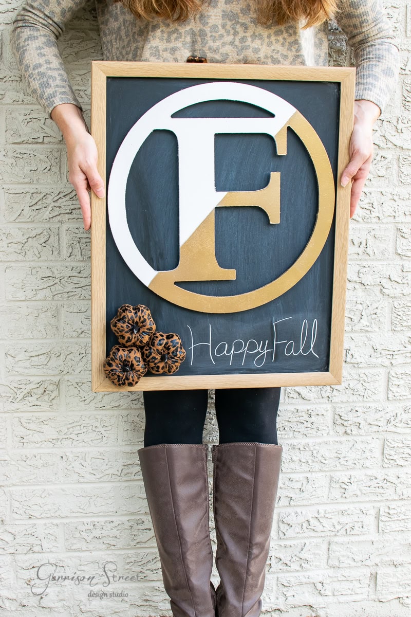 A person in brown boots and a leopard print top holds a DIY decorative sign featuring a large letter "F" and "Happy Fall" on a black backdrop, adorned with leopard print fabric flowers, resembling elements of an autumn wreath. The sign is charmingly displayed against a brick wall.