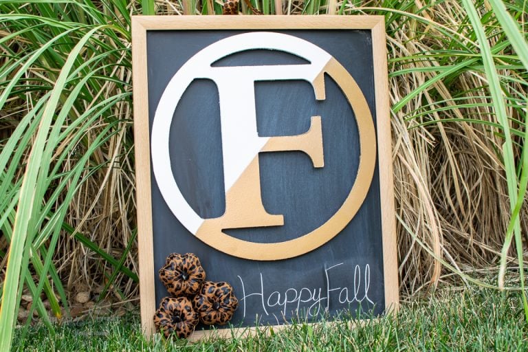 A decorative blackboard with a large, stylized letter "F" in the center, bordered by a wooden frame. This DIY board displays "Happy Fall" and is adorned with pinecones, echoing a charming wreath, all set against tall grass for an autumnal touch.