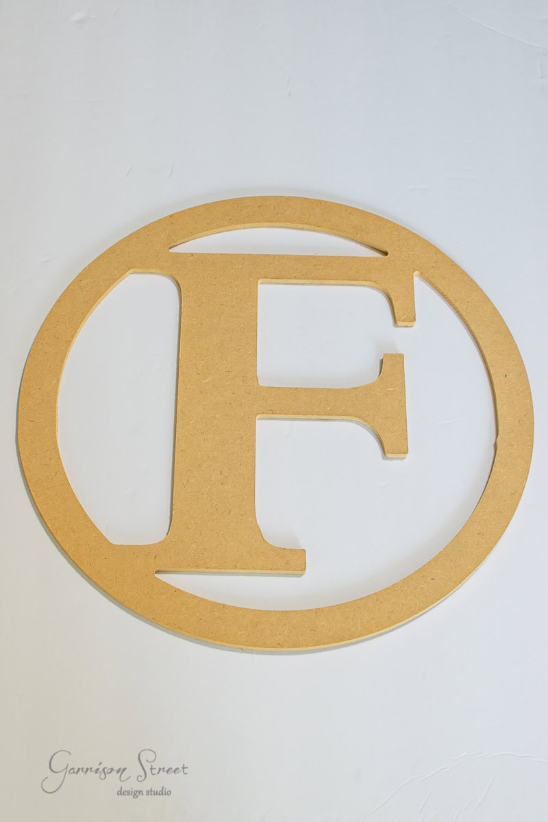 A wooden letter "F" is elegantly centered within an oval shape, reminiscent of a DIY wreath, resting on a plain white background.