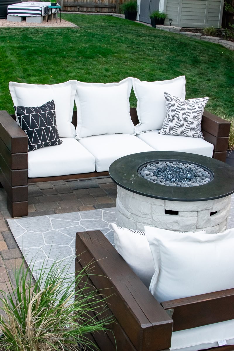 A cozy outdoor patio scene showcases a modern outdoor sofa set with white cushions and decorative black and white pillows. A round stone fire pit table is central, surrounded by green lawn and pavers, creating an inviting atmosphere.