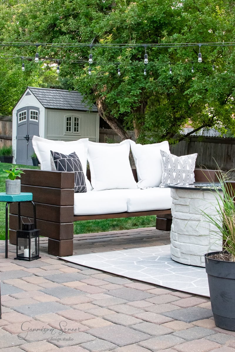 A cozy outdoor patio set with a modern wooden bench and an outdoor sofa, both adorned with white and patterned pillows, sits beside a round stone fire pit. String lights hang above, while a small shed with greenery surrounds the area in the background.