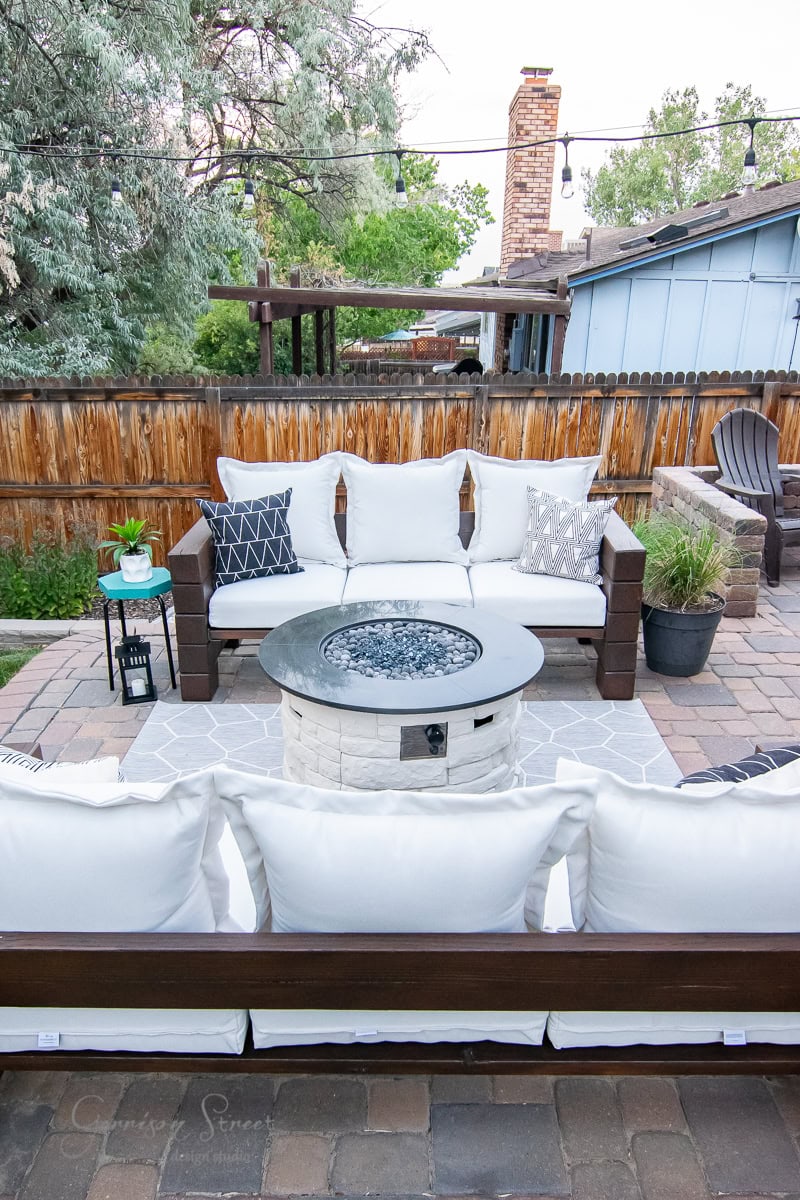 A cozy outdoor patio features an inviting circular fire pit table, encircled by wooden outdoor sofas with plush white cushions. Decorative pillows in various patterns add flair to the seating. A lantern sits on the left, with a wooden fence providing a charming backdrop.