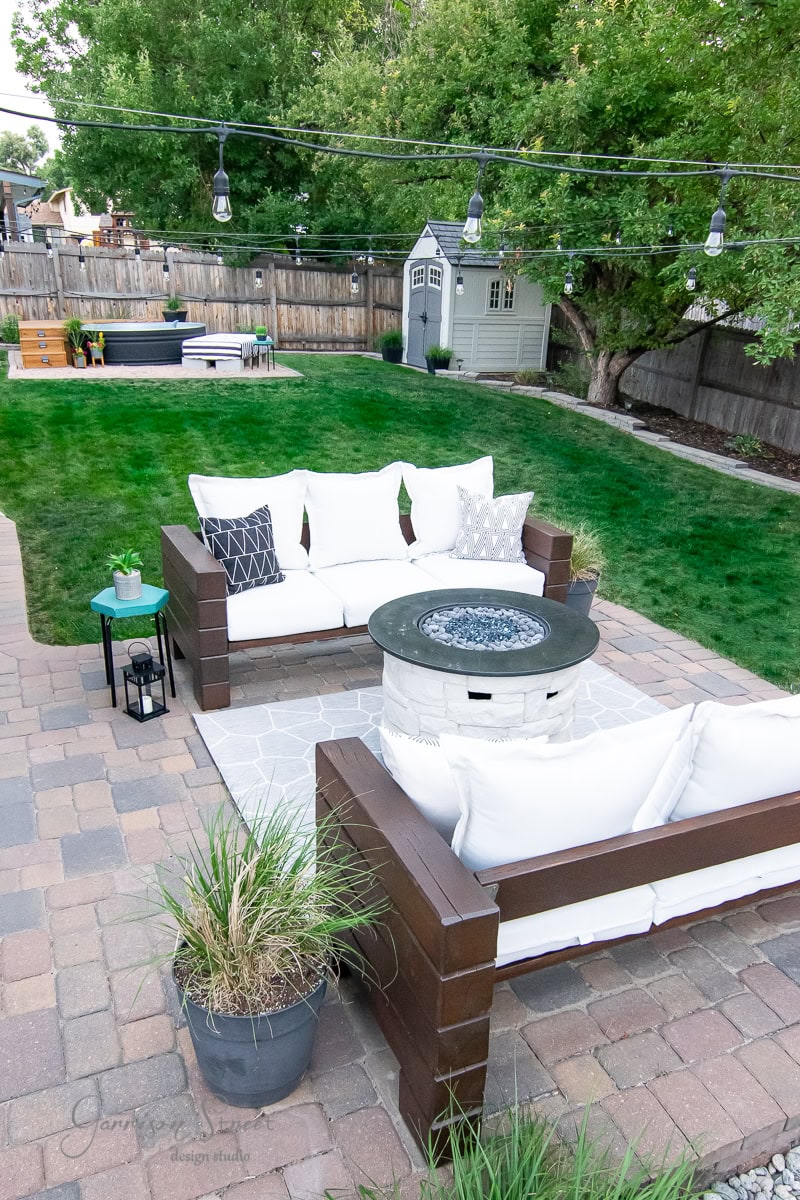 An outdoor patio showcases wooden couches with white cushions and an elegant outdoor sofa set around a round fire pit. String lights twinkle overhead, while a grassy lawn, trees, and a small shed create a serene backdrop. Paved flooring and potted plants enhance the cozy ambiance.