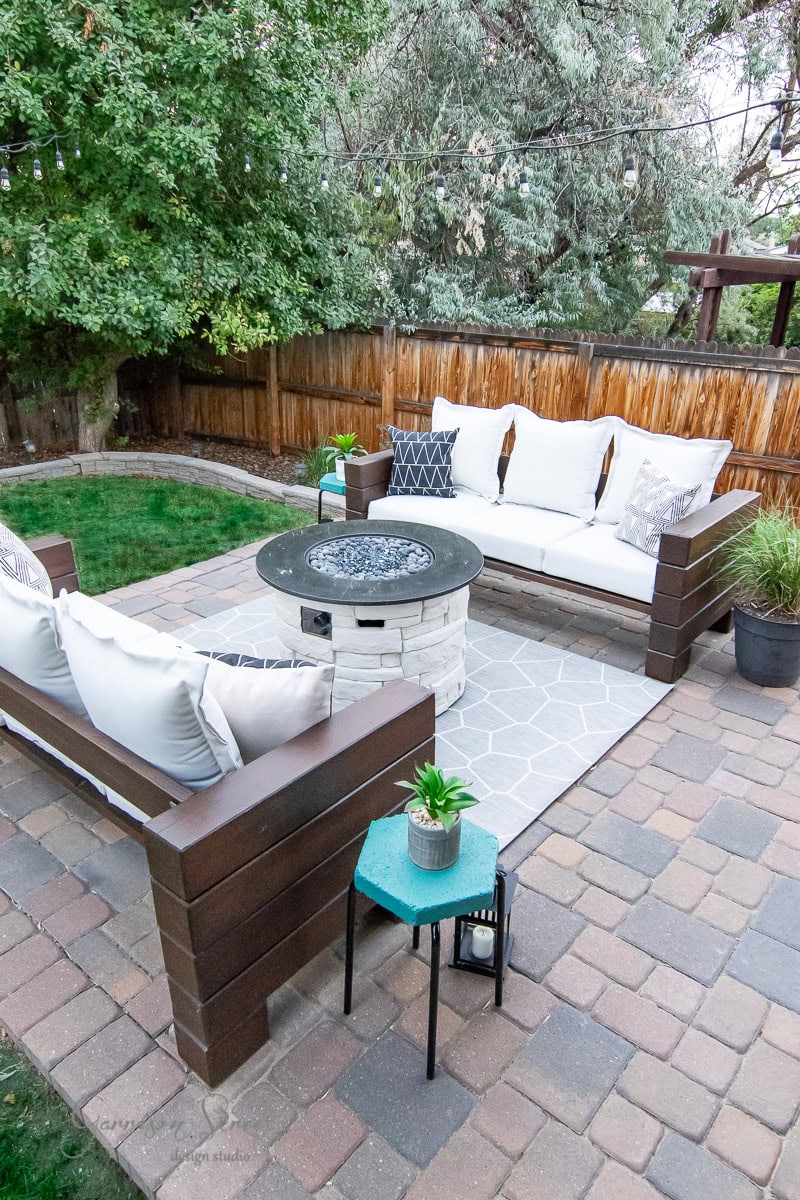 A cozy outdoor patio features two wooden outdoor sofas with white cushions around a circular fire pit table. A small teal side table with a plant sits nearby. The area is bordered by a wooden fence and lush green trees, creating an inviting and serene atmosphere.