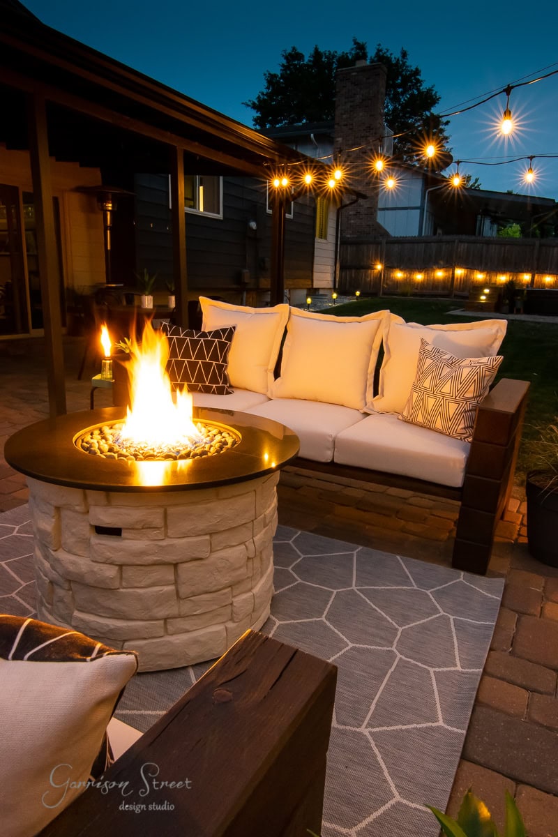 Cozy outdoor patio with a stone fire pit and a comfortable seating area featuring an inviting outdoor sofa adorned with cream and patterned pillows. Warm string lights hang overhead, creating a welcoming ambiance. A geometric rug lies beneath the furniture, set against the twilight sky.