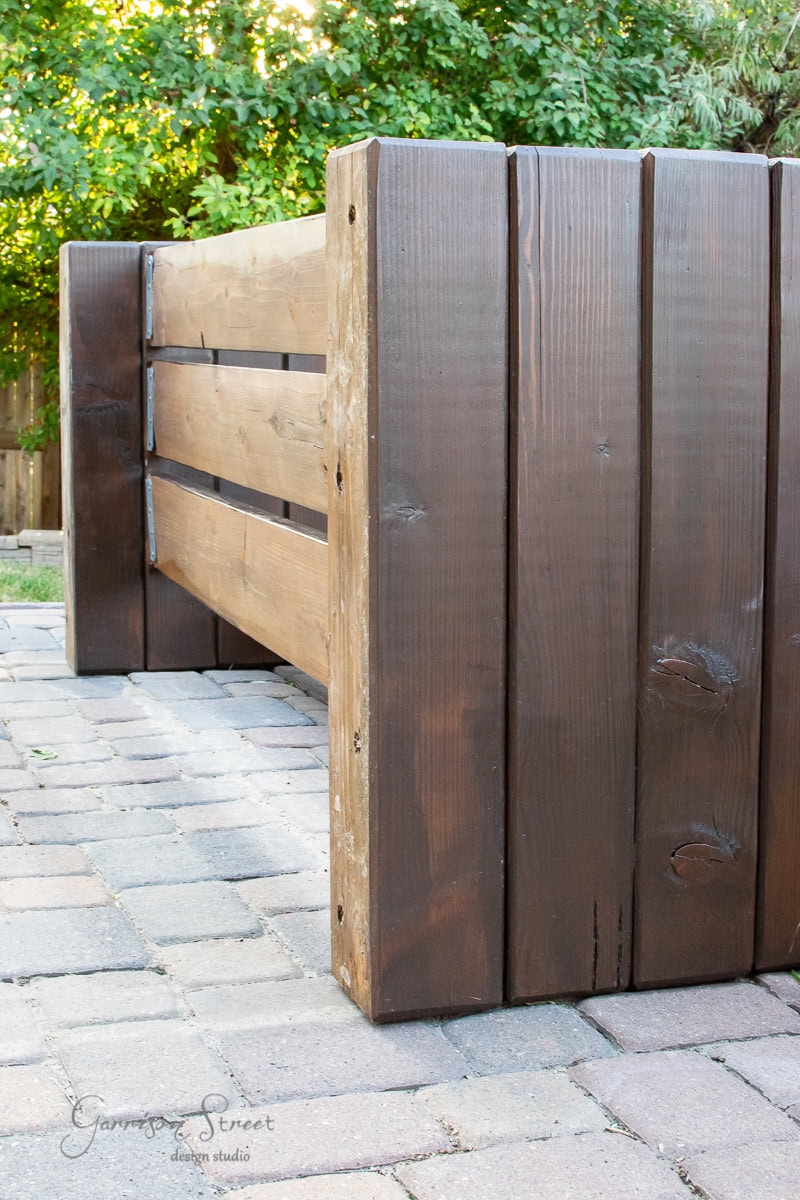 A wooden planter box with wide, dark-stained planks is placed on a stone-paved surface near an elegant outdoor sofa. Lush green foliage is visible in the background, enhancing the modern, rustic style.