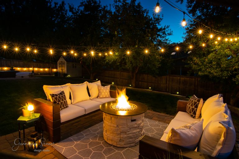 A cozy outdoor seating area at dusk features a circular stone fire pit surrounded by plush outdoor sofas. String lights hang overhead, casting a warm glow. Lush trees and a wooden fence frame the backyard, creating an inviting atmosphere.