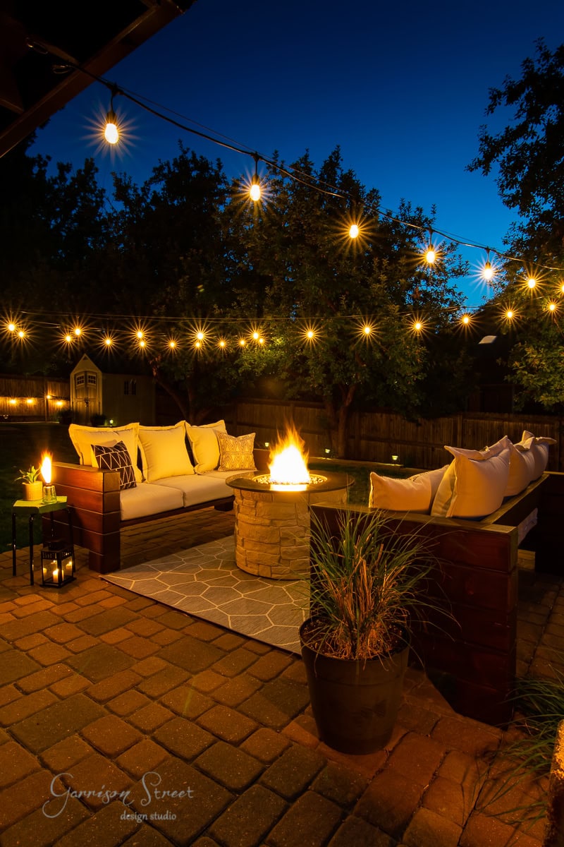 A cozy outdoor patio at dusk features string lights above, a fire pit glowing at the center, surrounded by cushioned seating and an inviting outdoor sofa. Potted plants and a lantern with a lit candle complete the charming atmosphere.