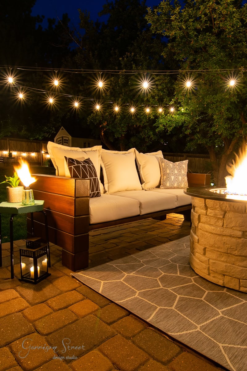 A cozy outdoor patio at night features an inviting outdoor sofa, a fire pit emitting a warm glow, string lights overhead, and a lantern on a small table beside the sofa. The area is set on a brick floor with a geometric patterned rug.