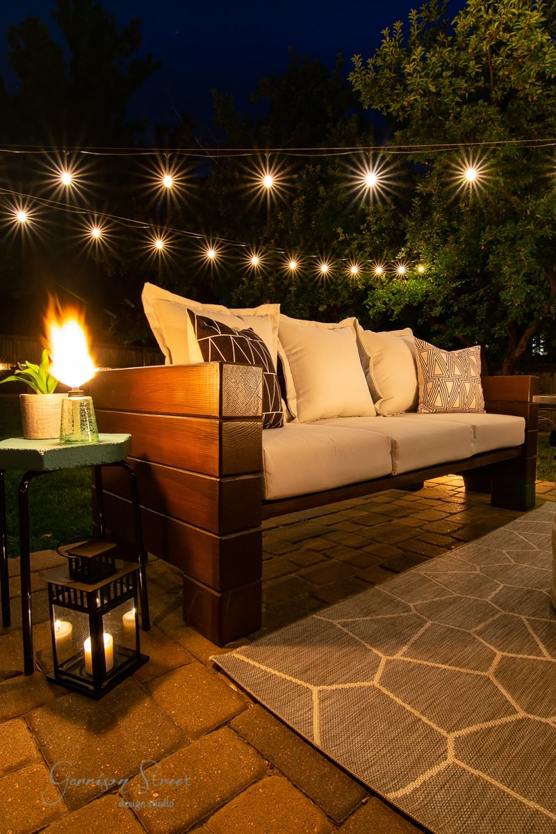 A cozy outdoor patio setting at night features an inviting wooden outdoor sofa adorned with white cushions and patterned pillows. String lights hang above, while a lantern glows on a side table next to a fire feature, creating a warm ambiance.