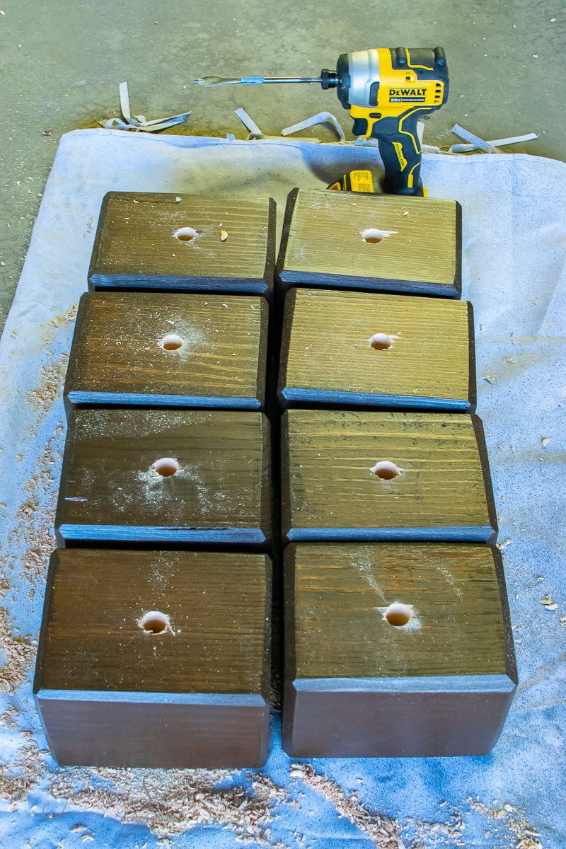 A collection of eight wooden blocks with drilled holes is arranged on a light blue cloth, resembling the foundation for an outdoor sofa. Wood shavings are scattered around, while a yellow and black power drill rests in the background.