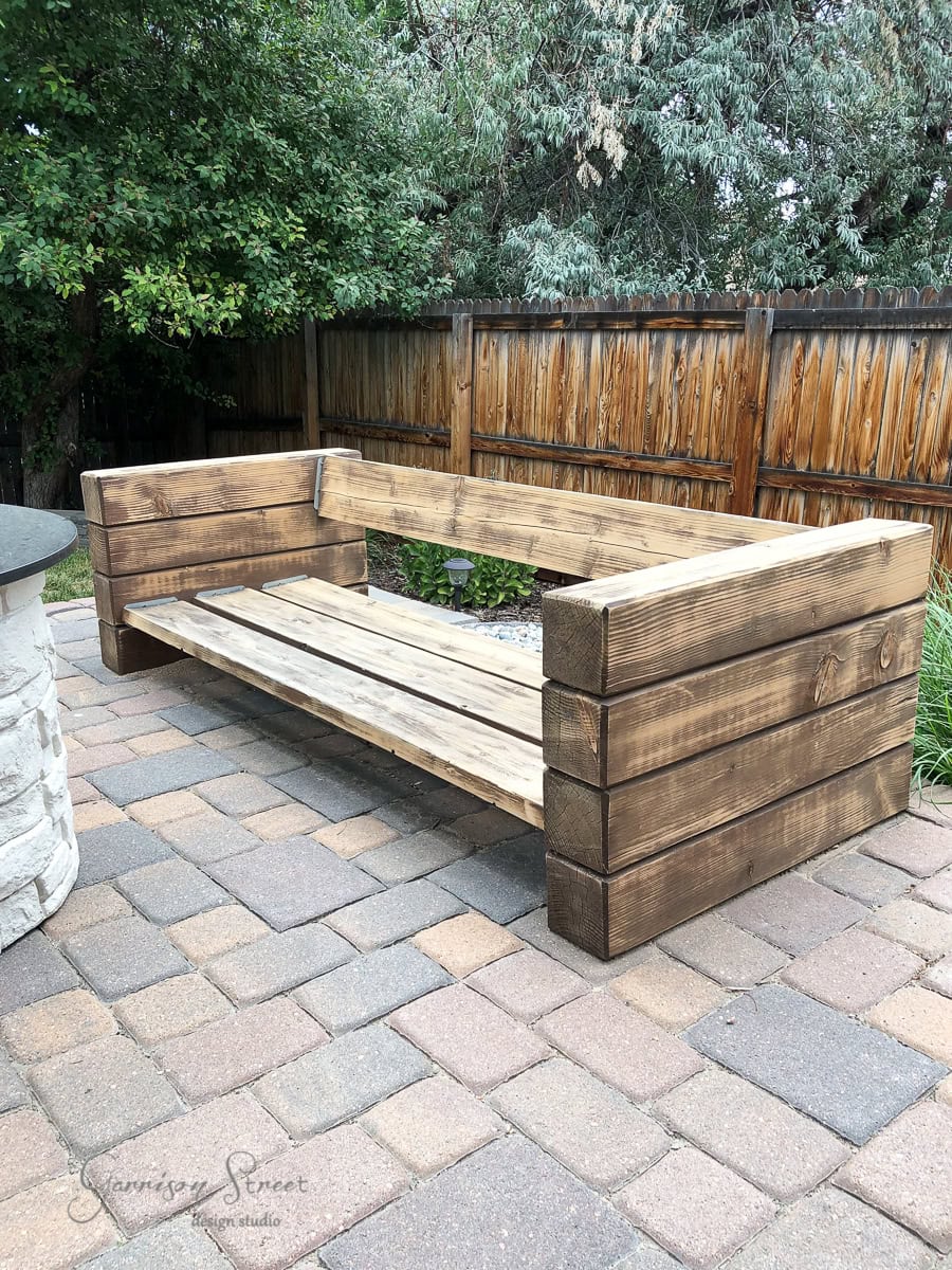 A wooden outdoor bench with wide, rustic planks creates a cozy seating area reminiscent of an outdoor sofa on a stone patio. The bench is set against a wooden fence with leafy greenery visible in the background.